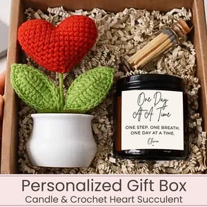 May include: A gift box containing a red crocheted heart on a green stem in a white ceramic pot, a glass vial of matches, and a black candle jar with a white label reading One Day At A Time, ONE STEP. ONE BREATH. ONE DAY AT A TIME. Olivia.