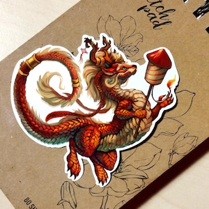 Puede incluir: Una pegatina con una ilustración detallada de un dragón rojo y dorado sosteniendo un fuegos artificiales. El dragón tiene un cuerpo largo y sinuoso y está sobre un fondo marrón con bocetos florales. El fuegos artificiales tiene una parte superior roja y una llama.