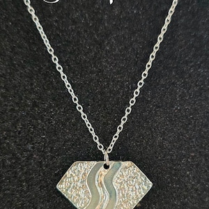 Könnte beinhalten: Eine silberplattierte Halskette mit einem diamantförmigen Anhänger mit einer strukturierten Oberfläche und einem wellenförmigen Design. Der Anhänger ist an einer silbernen Kette befestigt.