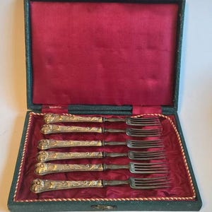 Peut inclure: Un ensemble de six fourchettes anciennes dans un coffret vert doublé de velours rouge. Les fourchettes ont des manches argentés ornés avec des détails dorés et des dents sombres. Le coffret est bordé d'une bordure décorative.