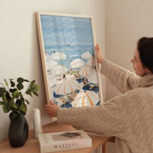 Beach scene featuring striped umbrellas and sunbathers by the calm ocean, painted in soft blue, peach, and cream tones — printable coastal wall art.
