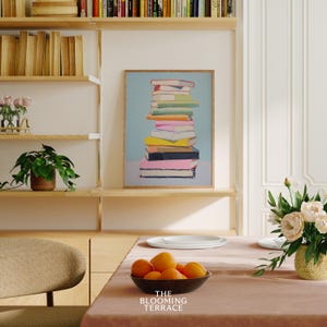 K&ouml;nnte beinhalten: Ein gerahmter Druck einer Stapel bunter B&uuml;cher auf hellblauem Hintergrund. Der Druck h&auml;ngt an einer Wand in einem Raum mit Holzregalen und einem Tisch mit einer Sch&uuml;ssel Orangen.