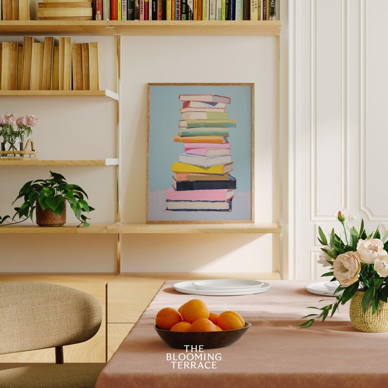 K&ouml;nnte beinhalten: Ein gerahmter Druck einer Stapel bunter B&uuml;cher auf hellblauem Hintergrund. Der Druck h&auml;ngt an einer Wand in einem Raum mit Holzregalen und einem Tisch mit einer Sch&uuml;ssel Orangen.