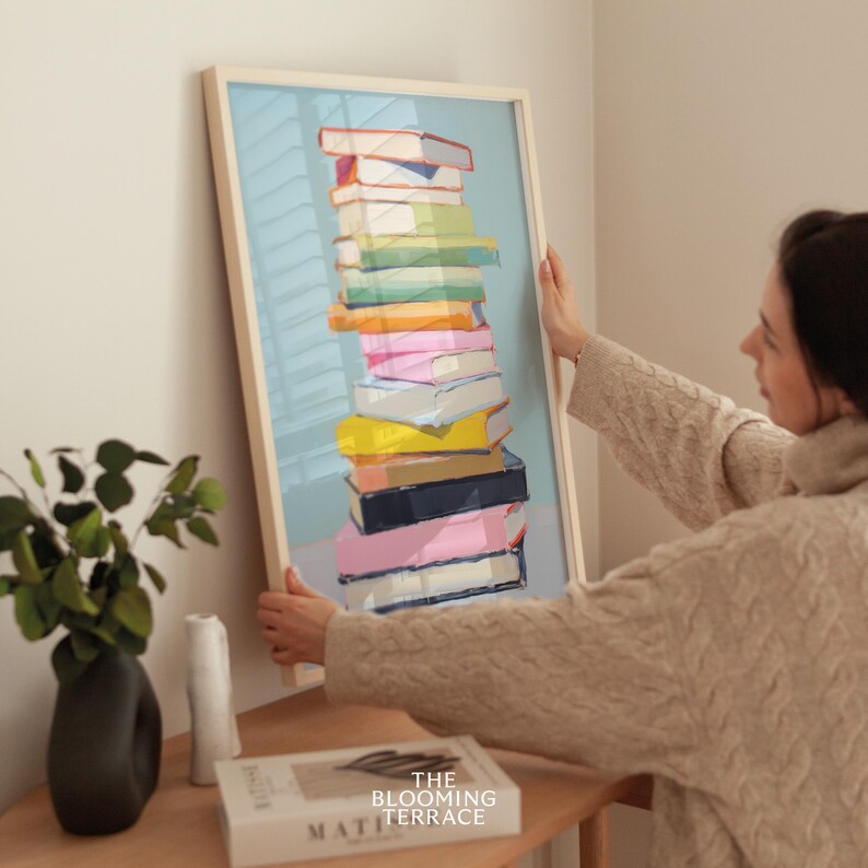K&ouml;nnte beinhalten: Ein gerahmter Druck einer Stapel bunter B&uuml;cher vor einem hellblauen Hintergrund. Der Druck ist in einem wei&szlig;en Rahmen und wird von einer Person an eine Wand geh&auml;ngt.