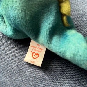 May include: A green and blue Ty Beanie Baby with a white tag that reads "The Beanie Babies Collection" and "Iggy".