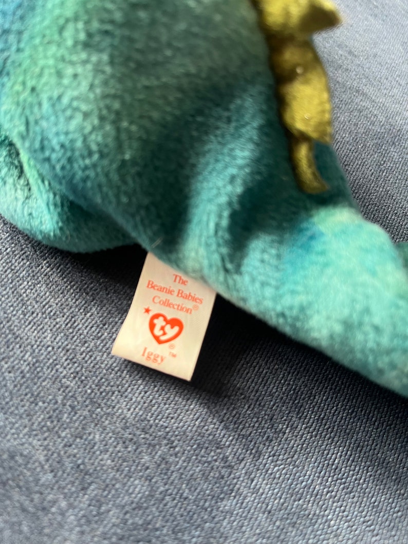 May include: A green and blue Ty Beanie Baby with a white tag that reads "The Beanie Babies Collection" and "Iggy".