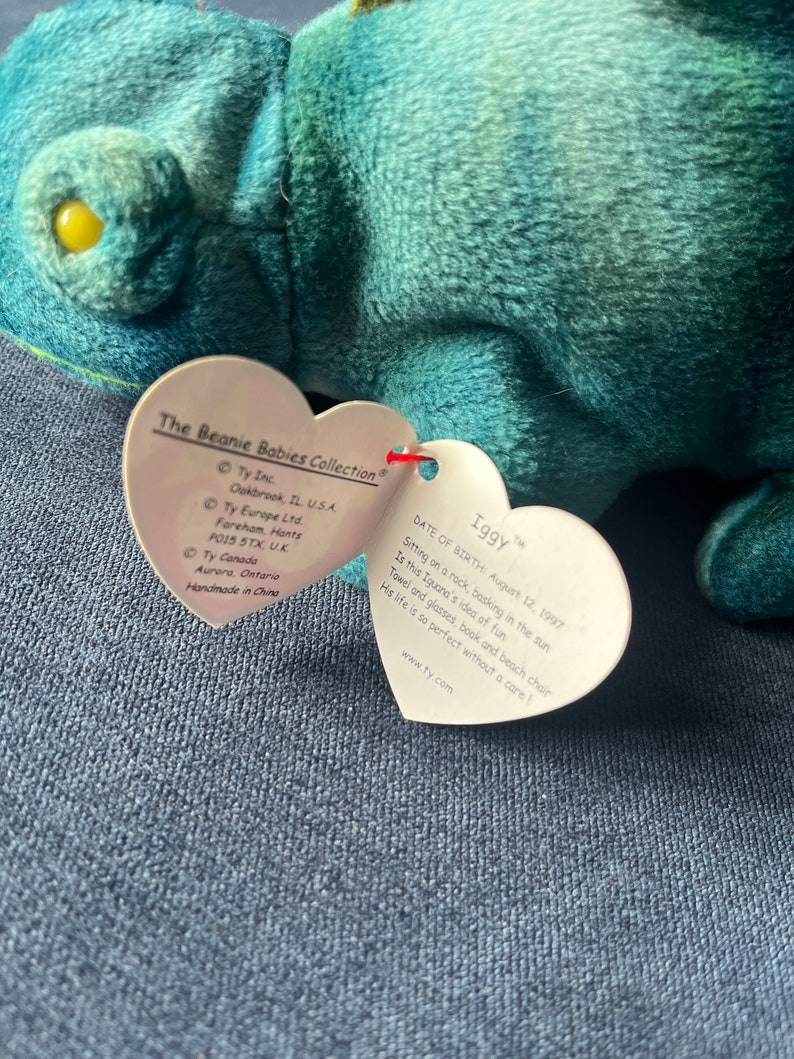 May include: A green and blue plush toy iguana named Iggy. The tag reads "Date of Birth: August 12, 1997" and describes Iggy sitting on a rock, basking in the sun. The tag also includes the text "www.ty.com".