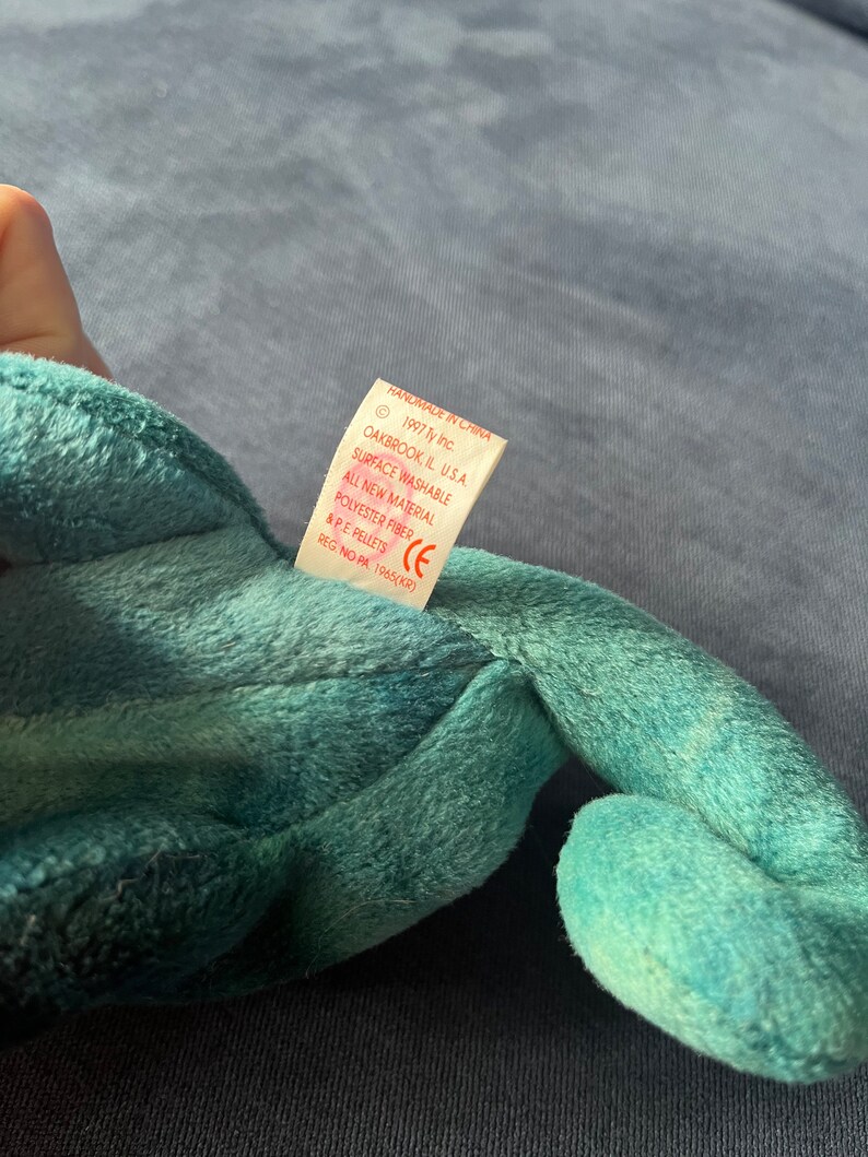 May include: A teal green beanie baby with a white tag that reads "Handmade in China", "1997 Ty Inc.", "Oakbrook, IL U.S.A.", "Surface Washable", "All New Material", "Polyester Fiber & PE Pellets", "CE", "Reg. No. PA 1965(KR)"