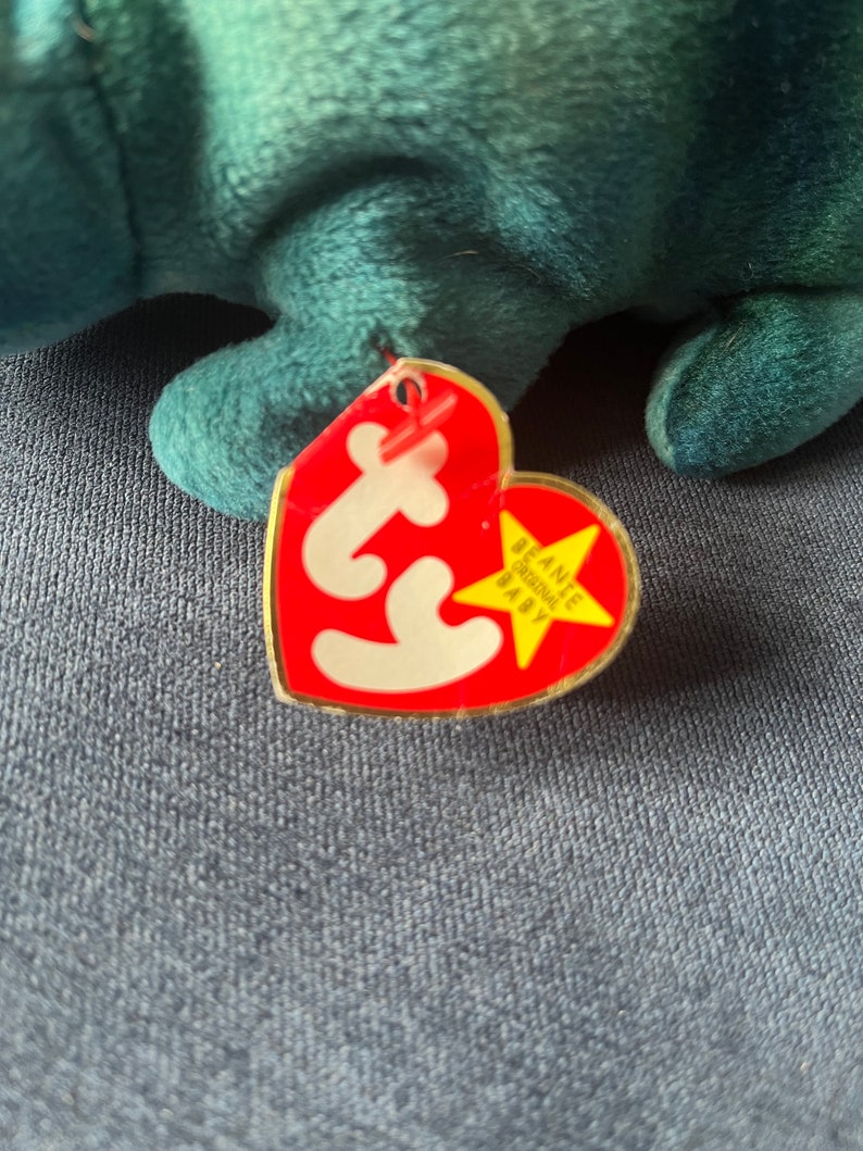 May include: Red Ty Beanie Baby tag with a white star and the text "Beanie Baby" in yellow. The tag is attached to a teal plush toy.