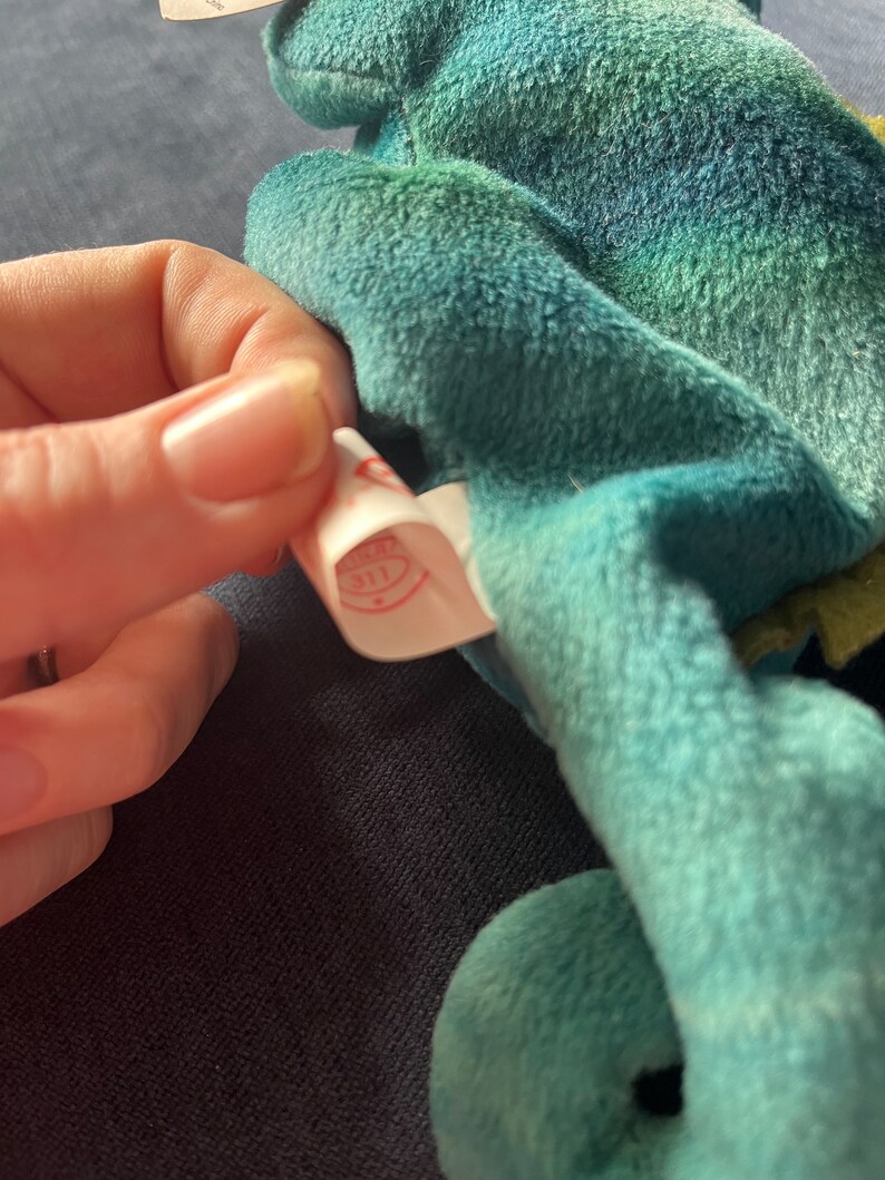 May include: A close-up of a teal blue plush toy with a white tag attached. The tag has a red circle with the word "Ty" inside.