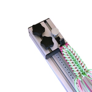 Könnte beinhalten: Ein silberner Metallwebstuhl mit zwei schwarzen sternförmigen Knöpfen und einem silbernen Knopf. Ein geflochtenes Band in Pink und Grün ist durch den Webstuhl gefädelt. Der Webstuhl hat ein Lineal mit den Zahlen 1 bis 13.