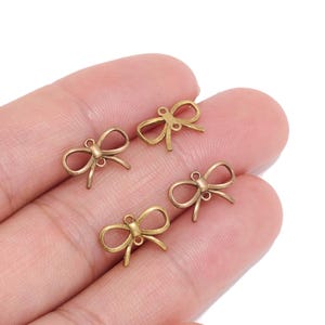Op de afbeelding: Vier kleine strikbedels van antiek messingkleurig metaal, elk met een delicaat lusontwerp en een enkele ronde bevestigingsring aan de bovenkant, gepresenteerd als individuele sieradenonderdelen voor knutsel- en decoratieve doeleinden.