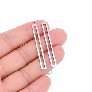 May include: Two silver-toned metal rectangular charms with an open centre design. Each charm features a thin, elongated frame with a small circular loop at the top for attachment, showcasing a smooth, reflective finish and a clean, geometric shape.