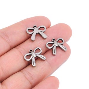 Op de afbeelding: Drie kleine, donkerkleurige metalen bedels in de vorm van een strik, elk met een gladde, gepolijste afwerking en een enkel rond oogje aan de bovenkant voor bevestiging. Deze sieradenonderdelen hebben een symmetrisch ontwerp met twee lussen.
