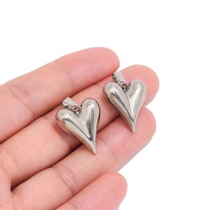 May include: Two polished silver-toned metal heart-shaped pendants, each featuring a smooth, rounded surface and a small integrated bail at the top for attachment. The pendants have a reflective finish and a distinct three-dimensional shape.