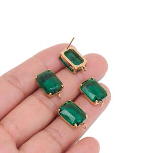 May include: Four rectangular emerald green glass charms set in gold-toned metal frames. Each charm features a faceted surface and a small metal loop at the bottom for attachment. One charm is positioned vertically with a visible metal post on the back.