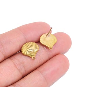 Könnte beinhalten: Zwei kleine muschelförmige Anhänger aus goldfarbenem Metall, jeweils mit einer texturierten, fächerförmigen Oberfläche mit vertikalen Rillen und einer kleinen kreisförmigen Öse an der Unterkante zur Befestigung, präsentiert als einzelne Schmuckkomponenten.