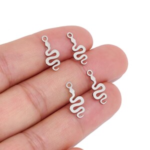 May include: Four small, silver-toned metal snake charms, each featuring a wavy, serpentine body and a circular loop at the top for attachment. The charms have a flat, smooth finish and are arranged in a cluster, showcasing their uniform design.