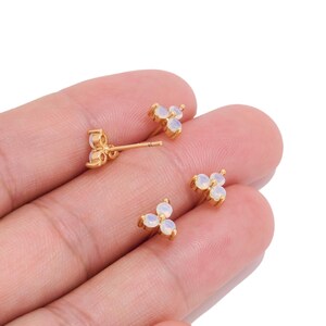 May include: Three gold stud earrings, each featuring a cluster of three small, round, iridescent white stones arranged in a triangular pattern. The earrings have a metallic gold finish and include a thin post and backing mechanism for attachment.