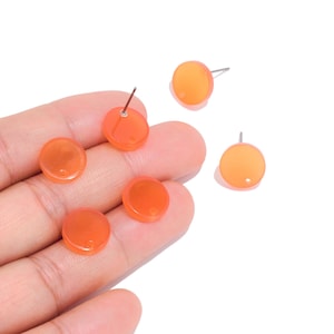 Pode incluir: Cinco brincos pequenos, redondos e de cor laranja translúcida, com uma superfície lisa e plana. Cada brinco possui um pino metálico fino fixado na parte de trás para fixação. Os brincos são circulares e têm uma cor laranja vibrante e sólida em todo o material.