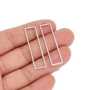 May include: Two silver-toned metal rectangular frame charms, each featuring a thin, open-centre design with a small circular loop at the top for attachment. These geometric components have a smooth, metallic finish and a clean, linear structure.