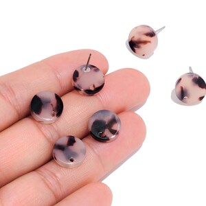 May include: Five small, circular stud earrings featuring a light pink and brown tortoiseshell pattern. Each earring has a smooth, flat surface with a metallic post attached to the back, and some pieces include a small hole near the edge.