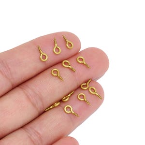 May include: A collection of small gold-toned metal screw eyes, each featuring a circular loop at the top and a threaded screw shank at the bottom, designed for attaching to various surfaces for hanging or connecting small items securely.
