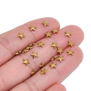 May include: A collection of small, metallic gold-colored star-shaped studs or embellishments. Each piece features a five-pointed design with a smooth, reflective surface. These tiny star components are scattered across the palm of a hand.