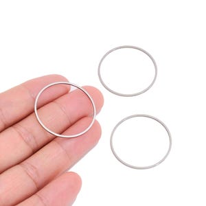 Puede incluir: Tres anillos metálicos circulares y delgados con un acabado plateado liso. Un anillo se encuentra en la palma de una mano, mientras que otros dos anillos idénticos están colocados por separado cerca. Estos aros metálicos ligeros tienen un diámetro uniforme.