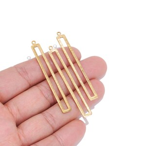 May include: Three elongated, rectangular gold-toned metal jewelry charms with a brushed finish. Each charm features a hollow center and a small circular loop at the top for attachment, arranged in a parallel formation against a plain background.