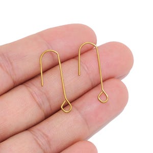 Puede incluir: Dos ganchos para pendientes de metal en tono dorado con un diseño curvo en forma de U. Cada gancho presenta un pequeño bucle circular en el extremo inferior para sujetar componentes de joyería, con un acabado de textura metálica mate.
