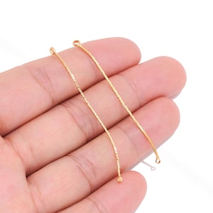 Puede incluir: Dos barras metálicas delgadas y curvas de tono dorado con una superficie texturizada y brillante. Cada barra presenta un pequeño bucle circular en ambos extremos, diseñado para componentes de joyería. El metal tiene un acabado brillante.