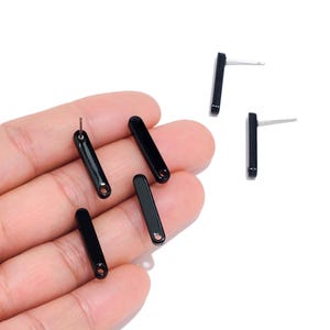 May include: A collection of small, glossy black bar-shaped stud earrings. Each earring features a smooth, elongated rectangular design with a rounded bottom edge and a small circular hole at the base, paired with a thin metal post for attachment.
