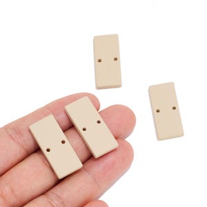 Puede incluir: Tres botones rectangulares de color beige, cada uno con dos pequeños agujeros centrados a lo largo. Los botones tienen una superficie lisa y plana con esquinas redondeadas, con dos piezas colocadas por separado y dos en una palma.