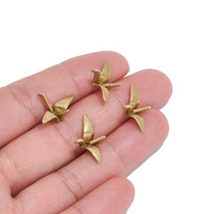 May include: Four small, metallic gold-toned charms shaped like origami cranes. Each charm features a smooth, polished surface with defined wings, a pointed beak, and a tail, capturing the geometric form of a folded paper bird in a miniature size.