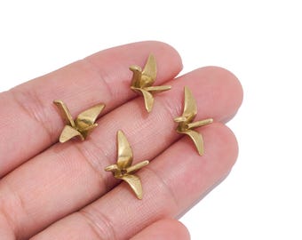May include: Four small, metallic gold-toned charms shaped like origami cranes. Each charm features a smooth, polished surface with defined wings, a pointed beak, and a tail, capturing the geometric form of a folded paper bird in a miniature size.