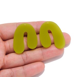 Puede incluir: Un par de pendientes de color verde oliva mate con una forma curva y arqueada. Cada pendiente tiene un pequeño orificio en la parte superior para su sujeción. Los pendientes presentan un acabado sólido y opaco con una silueta redondeada.