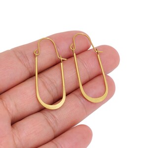 May include: A pair of gold-toned hoop earrings featuring an elongated, U-shaped design. The earrings have a smooth, metallic finish and a simple wire closure mechanism at the top, forming a continuous, minimalist oval silhouette for the earlobe.