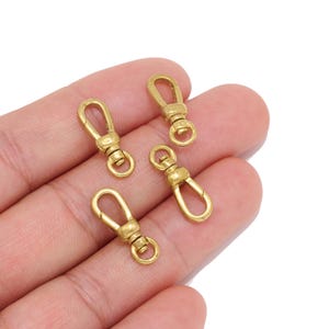 May include: Four gold-toned metal lobster claw clasps with a swivel base and a spring-loaded gate mechanism. These small hardware components feature a smooth, polished finish and are designed for connecting items such as jewelry or keychains.