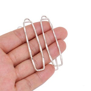 Op de afbeelding: Twee langwerpige, rechthoekige zilverkleurige metalen sieradenonderdelen met een gehamerd, getextureerd oppervlak. Elk stuk heeft een kleine ronde lus aan de bovenkant voor bevestiging, gekenmerkt door een slank, open frameontwerp met afgeronde hoeken.