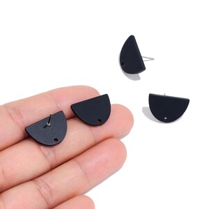 Puede incluir: Un conjunto de pendientes de botón en negro mate con forma de semicírculo. Cada pendiente presenta un diseño plano de media luna con un pequeño orificio circular en el borde inferior y un poste metálico para sujetarlo al lóbulo.