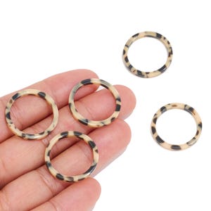 Può includere: Quattro anelli circolari con un motivo tartarugato beige e marrone. Due anelli sono posizionati su un palmo, mentre altri due anelli sono collocati separatamente nelle vicinanze. Ogni anello presenta una superficie liscia e arrotondata.