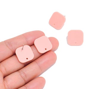 May include: Three light pink square earring components with rounded corners. Each piece features a small hole near the bottom edge and a metal post attached to the back, designed for jewelry assembly and crafting projects.