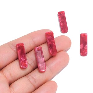 May include: Four rectangular acrylic pendants with rounded bottom edges, featuring a marbled pattern in shades of deep pink and red. Each piece has a small circular hole at the top for attachment, showcasing a glossy, smooth surface finish.