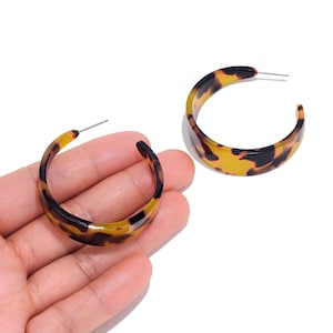 May include: A pair of hoop earrings featuring a glossy tortoiseshell pattern in shades of amber, brown, and black. The earrings have a wide, curved C-shape design with thin metal posts for attachment, showcasing a smooth, polished finish throughout.