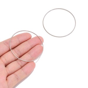 May include: Two thin, circular silver-toned metal rings with a smooth, plain surface. One ring is positioned flat against the palm of a hand, while the second identical ring is placed separately to the upper right on a plain white background.