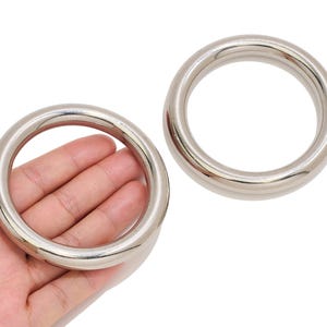 May include: Two circular metal rings with a smooth, polished silver-tone finish. Each ring features a thick, rounded cross-section and a seamless construction. The rings are solid, reflective, and have a uniform diameter throughout their structure.