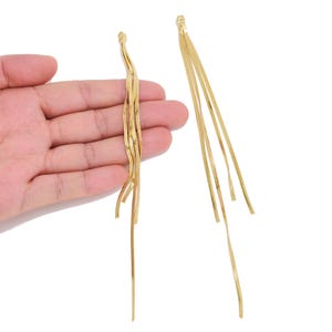 May include: A pair of long gold-toned dangle earrings featuring multiple thin, flat, flexible metallic strands that hang vertically from a single top attachment point, creating a fluid, cascading effect with a polished, reflective surface finish.