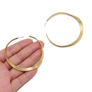 Op de afbeelding: Een paar grote goudkleurige oorringen met een plat, gedraaid ontwerp. De oorbellen hebben een gladde, metalen afwerking en een dunne stift sluiting. Eén ring rust op een handpalm, terwijl de andere er los naast is geplaatst.