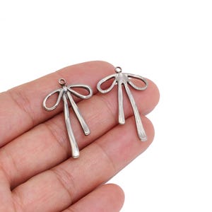 Op de afbeelding: Twee zilverkleurige metalen bedels in de vorm van strikken met lange, bungelende linten. Elke bedel is voorzien van een klein rond oogje aan de bovenkant voor bevestiging, met een gladde, reflecterende afwerking en een eenvoudig ontwerp.